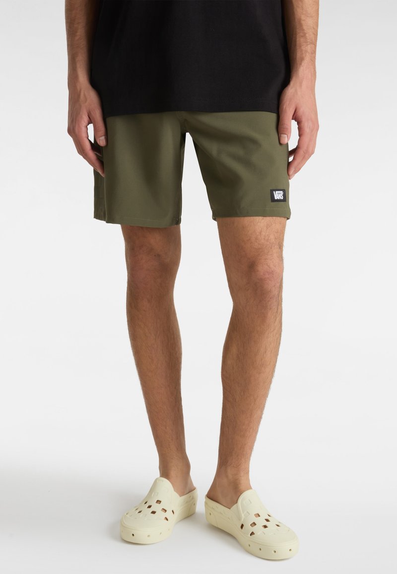 Shorts vert olive en tissu léger, arborant un écusson logo Vans, avec une coupe décontractée et droite. Portés avec des sabots beiges.