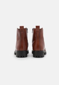 Paar braune Lederstiefeletten mit schwarzen klobigen Sohlen, sichtbaren hinteren Reißverschlüssen und Zuglaschen, von hinten auf weißem Hintergrund gezeigt.