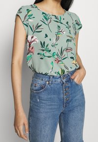 Blusa floreale di colore verde chiaro con maniche corte, caratterizzata da motivi rosa e verdi, abbinata a jeans in denim blu a vita alta con dettagli sui bottoni.