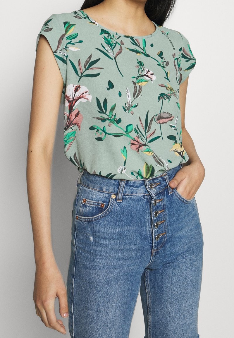 Blusa floreale di colore verde chiaro con maniche corte, caratterizzata da motivi rosa e verdi, abbinata a jeans in denim blu a vita alta con dettagli sui bottoni.