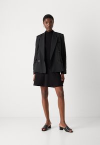 Blazer noir croisé sur un col roulé noir, associé à une mini-jupe noire et des mules à talons ouverts noires. Textures lisses.