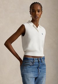 Kvinne med fletter i håret, iført en hvit ermeløs polotrøye og blå jeans, stående med en hånd i lomma mot en ensfarget bakgrunn.