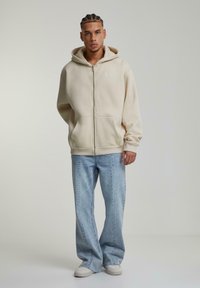 Felpa beige con zip e tasca frontale, abbinata a jeans a zampa d'elefante azzurri chiari e sneakers bianche. Texture liscia, vestibilità rilassata.
