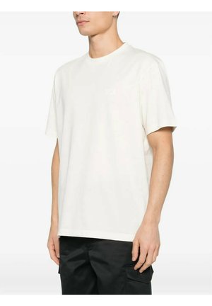 Uomo che indossa una t-shirt bianca semplice a maniche corte con scollo rotondo e pantaloni cargo neri, in piedi contro uno sfondo bianco.