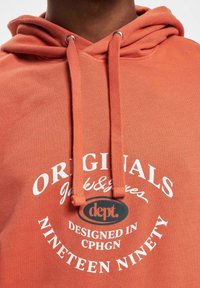 Hoodie in Lachsfarbe mit strukturiertem Gewebe, featuring ein gedrucktes Logo und Text in Weiß. Enthält eine Kordelzugkapuze und silberne Ösen.