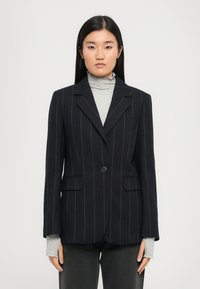 Navy pinstripe bleiseri yksinkertaisella napilla, lovetilla kauluksella ja kahdella etutaskulla, päällystettynä vaaleanharmaalla poolopaitalla.