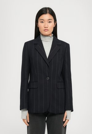 Blazer à rayures verticales bleu marine avec un bouton unique, revers crantés et deux poches avant, superposé à un col roulé gris clair.