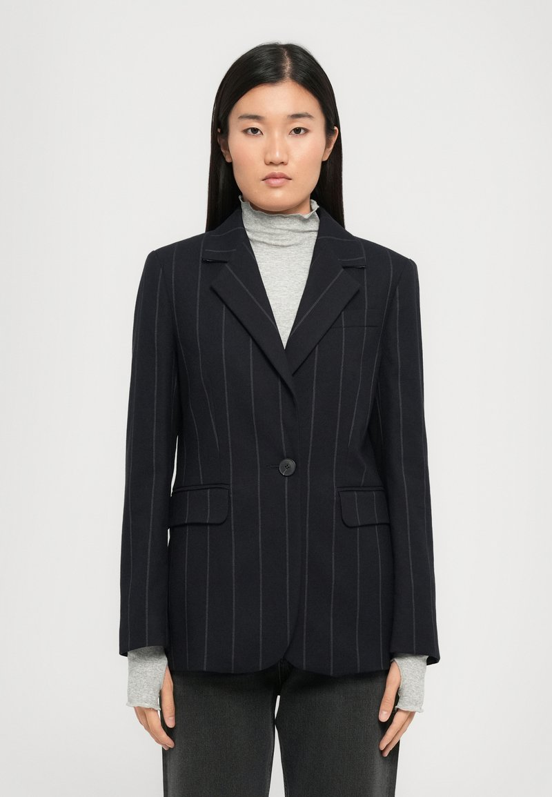 Navy pinstripe bleiseri yksinkertaisella napilla, lovetilla kauluksella ja kahdella etutaskulla, päällystettynä vaaleanharmaalla poolopaitalla.
