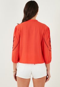 Rote, leichte Bluse mit dreiviertel Puffärmeln. Verfügt über schwarze gestickte Muster an den Ärmeln. Kombiniert mit weißen Shorts.
