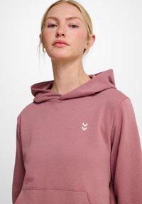 Rosa Kapuzenpullover mit einer Vorderseite Tasche, gefertigt aus weichem Stoff; hat ein dezentes cremefarbenes Chevron-Logo auf der linken Seite.