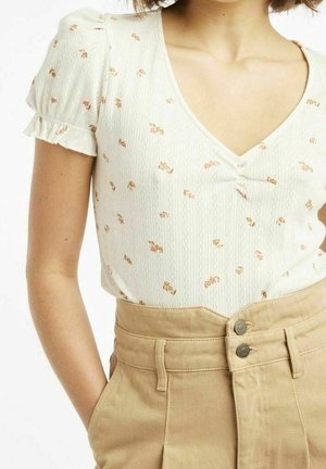 Vrouw draagt een wit topje met korte mouwen met kleine bloemmotieven en beige broek met hoge taille en dubbele knoopsluiting.