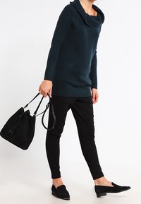 Sweter z dekoltem typu cowl, o strukturze prążkowanej, w ciemnozielonym kolorze, zestawiony z czarnymi spodniami slim fit i czarnymi balerinami, trzymając czarną torebkę na długich uchwytach.