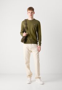 Olivgrünes Langarmhemd, cremefarbene Hose, weiße Sneaker und eine Schultertasche mit einem Logo-Riemen. Einfaches, modernes Design.