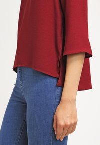 Top borgogna con una vestibilità rilassata, maniche corte arrotolate e fessure laterali, abbinato a jeans blu aderenti. Tessuto liscio e taglio casual.