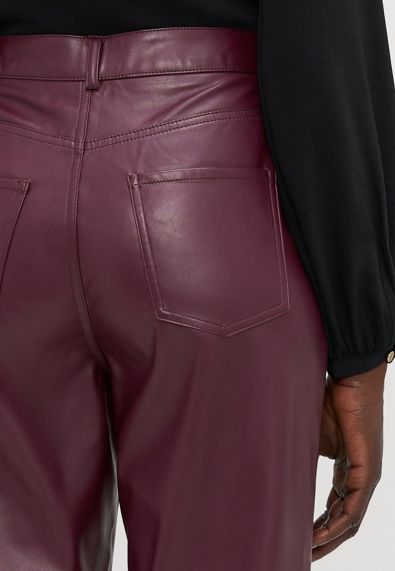 Pantaloni in finta pelle bordeaux con vita alta, dettagli sulle cuciture e due tasche posteriori, visti da dietro con una manica di una maglietta nera visibile.