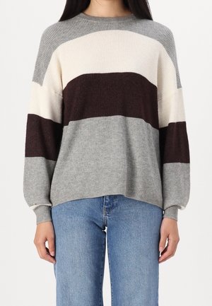 Femme portant un pull en maille côtelée avec des rayures horizontales gris, blanc et brun foncé, et un jean bleu, debout devant un fond blanc.
