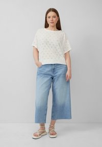 Witte oogjes top met korte mouwen, gecombineerd met lichtblauwe wijde jeans. Model draagt witte sandalen met bandjes. Eenvoudige, casual stijl.