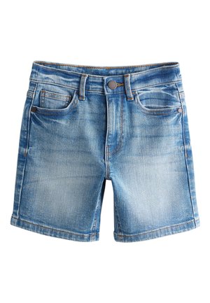 Dreng står og kigger til siden iført en ensfarvet hvid t-shirt, denimshorts, hvide sokker og sorte sneakers mod en neutral baggrund.