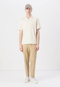 Ανοιχτό μπεζ τίτσιν της κοντομάνικης μπλούζας πόλο, εφαρμοστό, με V-λαιμό. Συνδυασμένο με ανοιχτό μπεζ παντελόνι σε στυλ tapered και λευκά αθλητικά παπούτσια, συμπληρωμένο με ένα ανοιχτό καπέλο.