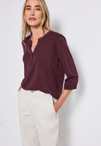 Chemise henley bordeaux avec un plastron à boutons et des manches 3/4, associée à un pantalon de couleur claire. Tissu doux et coupe décontractée.