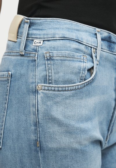 Κοντινό πλάνο τσέπης ανοικτού μπλε τζην με μικρή λευκή υφασμάτινη ετικέτα και ασημένιες πριτσίνια, φορεμένη με μαύρο τοπ.