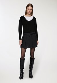 Giovane donna che indossa un maglione nero a V sopra una maglietta bianca, una gonna plissettata a righe con cintura, calze nere trasparenti e stivali neri al ginocchio.