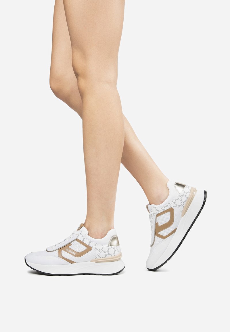Gambe che indossano sneakers bianche e beige con suole spesse, un piede piatto, l'altro sollevato sulle punte su uno sfondo bianco semplice.