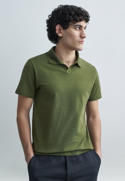 Polo a maniche corte verde oliva con colletto dritto, realizzato in tessuto testurizzato, mostrato con le mani nelle tasche, abbinato a pantaloni scuri.