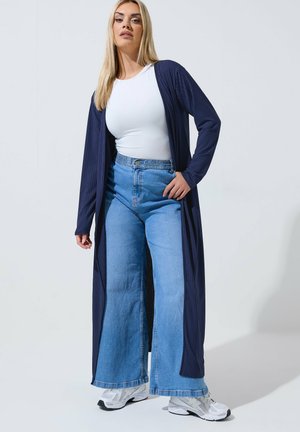 Kobieta stojąca w białym topie, szerokich niebieskich dżinsach, granatowym długim kardiganie oraz białych trampkach na tle jednolitego szarego tła.