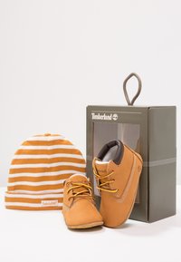 Brązowe buty Timberland z jasnobrązowej skóry z żółtymi sznurówkami, w zestawie z pasiastą, pomarańczowo-białą dzianą czapką, wyeksponowane w szarym pudełku.