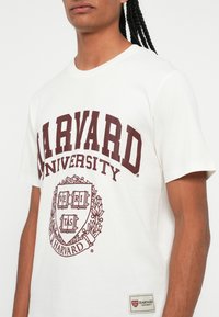 Tricou alb din bumbac cu imprimeu și emblemă maroniu "HARVARD UNIVERSITY" cu design floral pe piept. Eticheta este vizibilă pe partea inferioară.