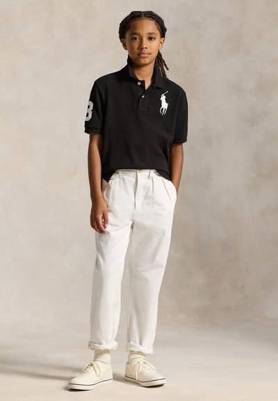 Lapsi, jolla on punotut hiukset, musta poolopaita, valkoiset housut ja valkoiset lenkkarit, seisoo neutraalin taustan edessä.