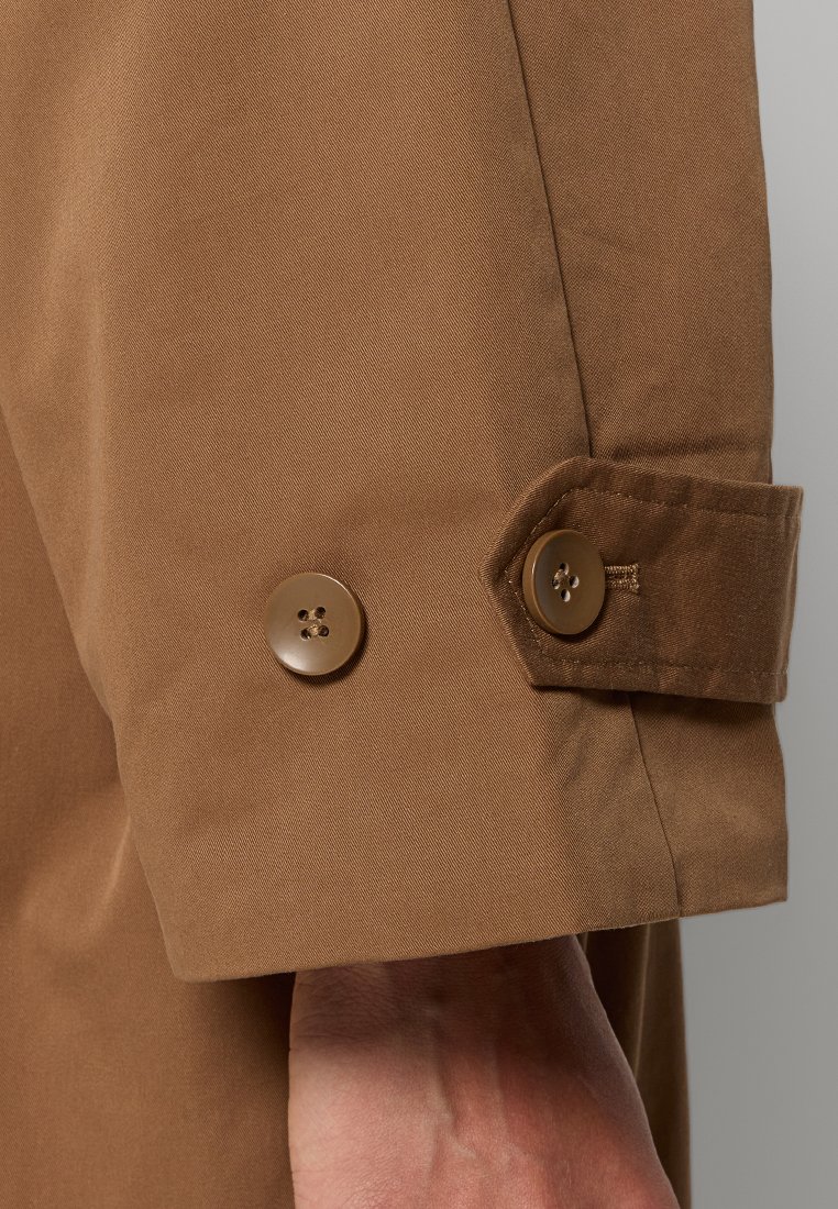 Manche de manteau marron avec deux boutons assortis et un détail de sangle, portée sur le poignet et la main d'une personne.