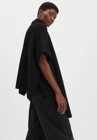 Donna con capelli corti che indossa un poncho di maglia nero largo e pantaloni neri, in posa contro uno sfondo bianco semplice.