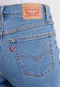 Vaqueros de mezclilla azul claro con un parche de cuero marrón que muestra el logo de "Levi Strauss & Co.", costuras naranjas y una pequeña etiqueta roja en el bolsillo trasero.