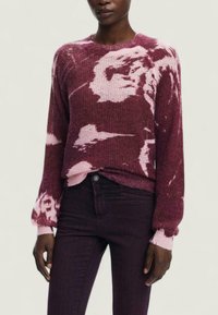 Sweter dzianinowy w burgundowym kolorze z różowymi abstrakcyjnymi wzorami. Ma okrągły dekolt i ściągacze przy mankietach. Połączony z ciemnofioletowymi dżinsami.