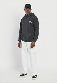 Sort hættetrøje med lynlås og frontlomme, hvid grafisk tekst. Sammen med hvide bukser og sorte sneakers med hvide såler. Sort kasket på hovedet.