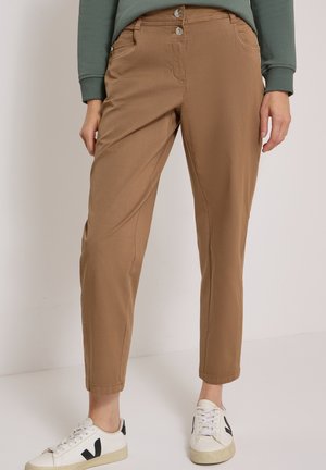 Femme portant un pantalon fuselé marron clair, un haut vert à manches longues et des baskets blanches avec des touches noires, debout sur un fond uni.