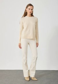 Γυναίκα που στέκεται σε εσωτερικό χώρο, φορώντας ένα κρεμ πλεκτό πουλόβερ, ταιριαστά κρεμ παντελόνια, μπεζ μποτάκια με μυτερή μύτη και χρυσά κρίκους σκουλαρίκια.