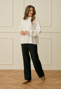 Ženska stoji na lesenem tleh, oblečena v belo bluzo z naborki, črne široke hlače in rjave čevlje z nizko peto, z rokami sklenjenimi skupaj.