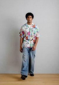 Άνδρας περπατά φορώντας πολύχρωμο πουκάμισο με κοντά μανίκια και κουμπιά, ανοιχτόχρωμο τζιν με φαρδιά μπατζάκια και μαύρα παπούτσια μπροστά σε έναν απλό τοίχο.