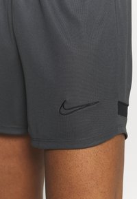 Šedé sportovní šortky s hladkou texturou, s černým logem Nike a malou boční kapsou. Design zahrnuje šité švy.