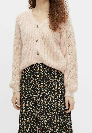 Femme portant un cardigan en grosse maille rose pâle avec des boutons et une jupe midi noire à fleurs sur un fond uni.