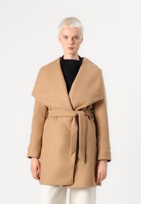 Manteau en laine marron avec un large col superposé, une taille ceinturée et des poches latérales. Le tissu semble doux et texturé.