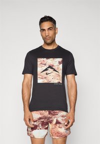 Svart t-skjorte med et frontgrafikk med et fjellmotiv og "Nike Trail," kombinert med røde og beige shorts med abstrakt mønster.