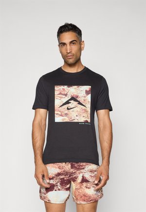 T-shirt noire avec un graphique sur le devant représentant un design de montagne et "Nike Trail", associé à un short à motifs abstraits rouge et beige.