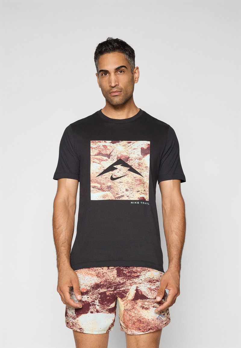 Svart t-skjorte med et frontgrafikk med et fjellmotiv og "Nike Trail," kombinert med røde og beige shorts med abstrakt mønster.