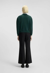 Una persona con capelli corti biondi sta in piedi di spalle indossando un maglione verde scuro, pantaloni a gamba larga neri e scarpe con plateau nere.
