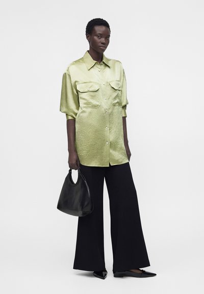 Γυναίκα που φοράει ένα ανοιχτό πράσινο φαρδύ σατέν πουκάμισο, μαύρο παντελόνι με φαρδιά πόδια, μαύρα μυτερά φλατ παπούτσια και κρατάει μια μαύρη τσάντα.