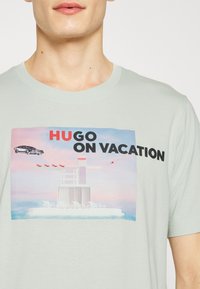 Camiseta verde claro con un estampado gráfico que presenta un coche, un hotel y el texto "HUGO DE VACACIONES". Hecha de algodón suave y transpirable.